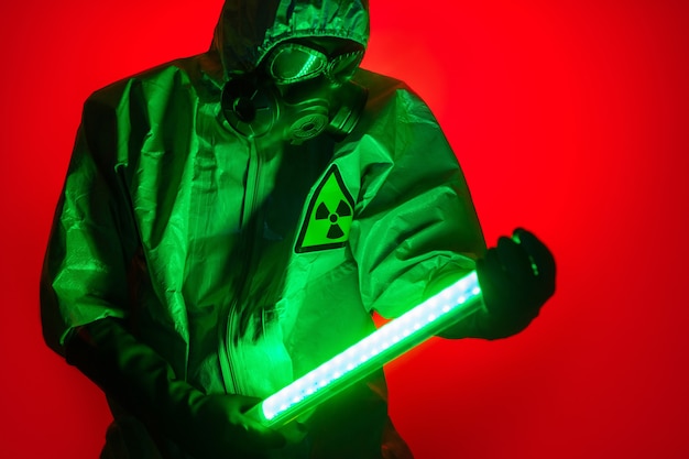 A man poses in a yellow protective suit with a hood on his head, with a protective gas mask, posing while standing against a red background, holding a green light uranium lamp in his hands.