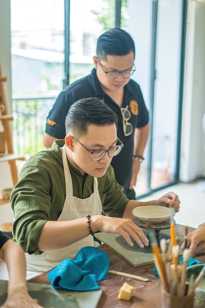 Man potter working on potters wheel making ceramic pot from clay in pottery workshop art concept Focus hand young man attaching clay product part to future ceramic product Pottery workshop