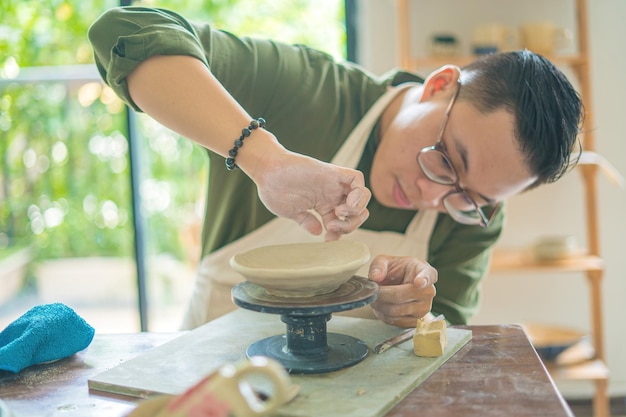 Man potter working on potters wheel making ceramic pot from clay in pottery workshop art concept Focus hand young man attaching clay product part to future ceramic product Pottery workshop