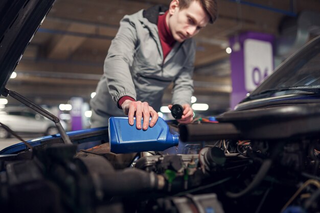 写真 開いたボンネットの車の青いボトルにオイルを注ぐ男