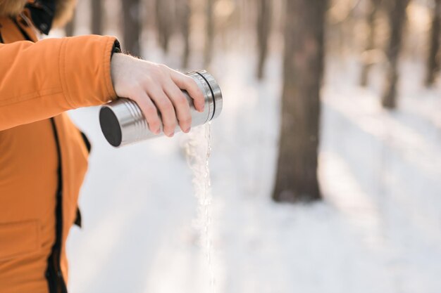 Man pours hot water from a thermos into a snow walking in snowy frozen winter forest at sunset