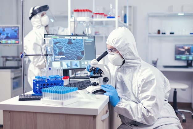Uomo in attrezzatura dpi che conduce indagini sul coronavirus guardando attraverso le indagini. scienziato in tuta protettiva seduto sul posto di lavoro utilizzando la moderna tecnologia medica durante l'epidemia globale.