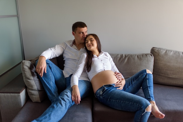 man and a pregnant woman are sitting hugging on the couch.