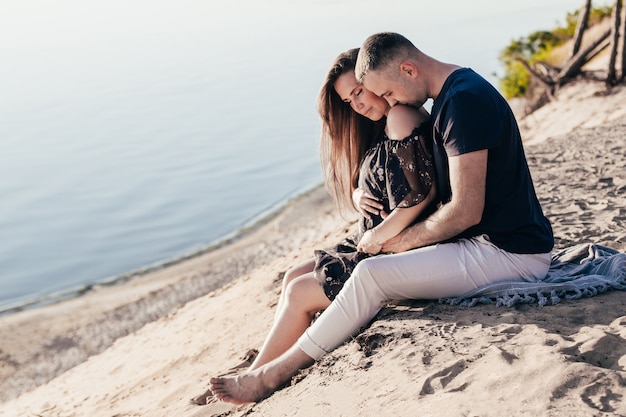 Man and a pregnant woman in an embrace on a sandy beach in summer at sunset