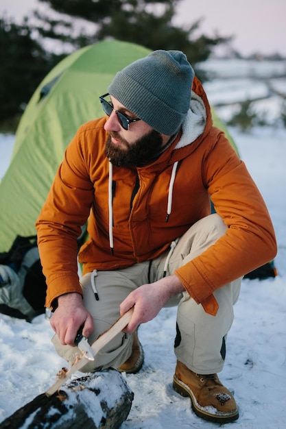 A man prepares wood wool with a knife for starting a fire in a winter forest at sunset Winter survival concept