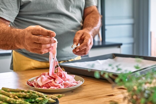 Photo man preparing young green asparagus sprouts wrapped in bacon on wooden table in the kitchen mans hand wrapping asparagus in ham for baking     person