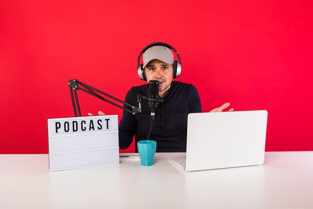 Man presenter with cap in podcast radio recording studio making hand gestures, next to a computer, a microphone and a light box with the word podcast, on red background. Podcasting, broadcast concept