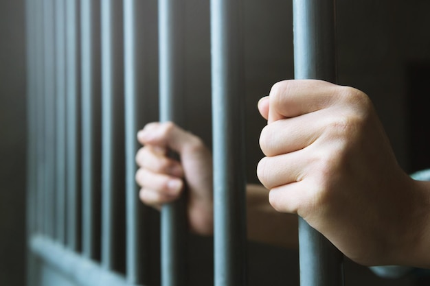 Man in prison hands of behind hold Steel cage jail bars offender criminal locked in jail
