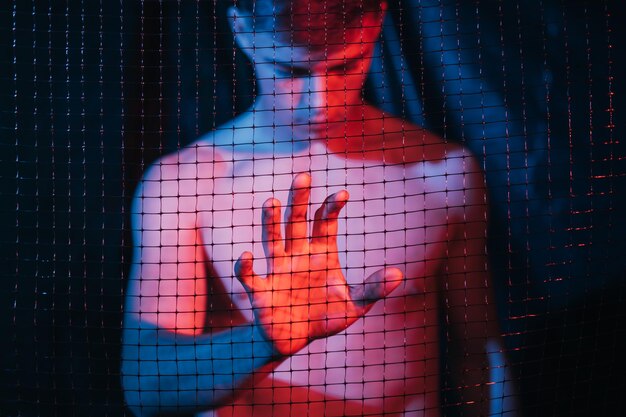 Man problem. Social inequality. Protection safety. Defocused depressed jobless shirtless guy touching mesh fence with hand in red blue neon light on blur dark background out of focus.