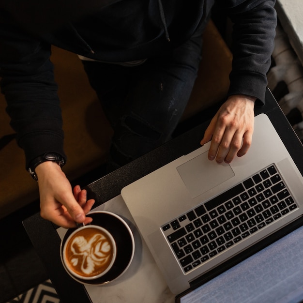 Man professional in black clothes sits in a cafe, drinks coffee and works on laptop, top view