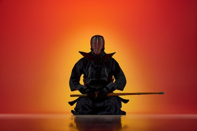 写真 グラデーション赤に対して竹刀でポーズをとって座っている制服を着た男プロ剣道選手