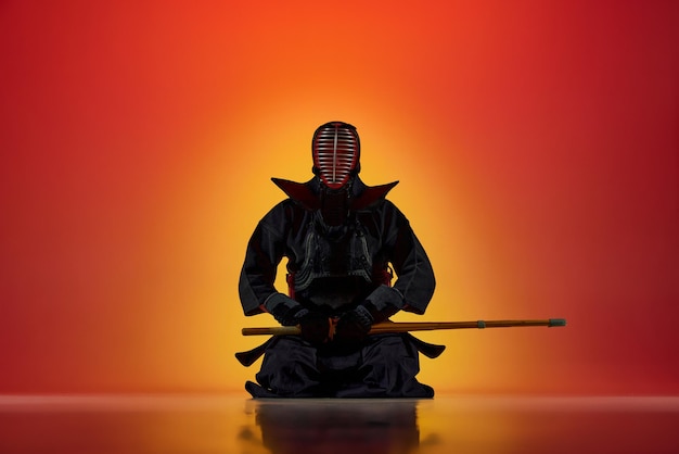 Man professional kendo athlete in uniform sitting posing with shinai sword against gradient red