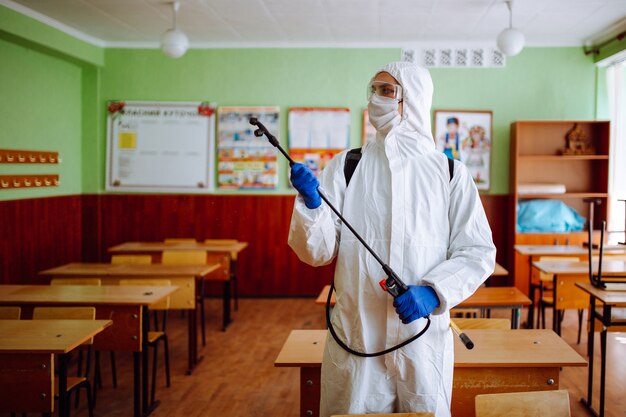 A man in protective antibacterial suit cleaning up the classroom with a spray with sanitizing liquid. Professional sanitary worker disinfects the auditorium with special equipment. Healthcare concept.