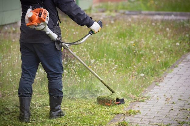 A man in protective clothing gloves and rubber boots mows the grass with a trimmer