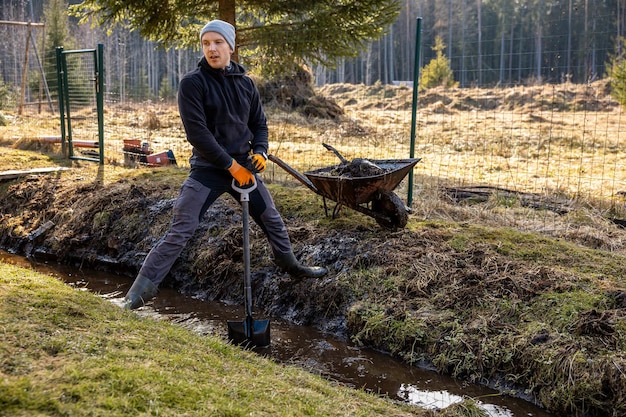 Человек в защитном снаряжении неустанно работает, чтобы выкопать грязный траншею с лопатой и тележкой на заднем дворе в начале весны
