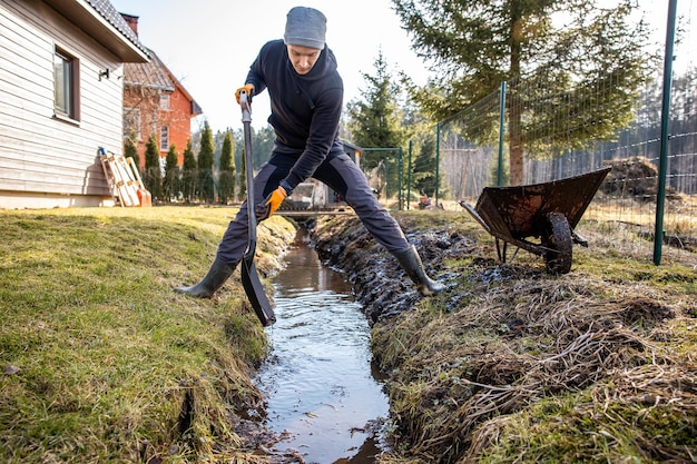 Человек в защитном снаряжении неустанно работает, чтобы выкопать грязный траншею с лопатой и тележкой на заднем дворе в начале весны