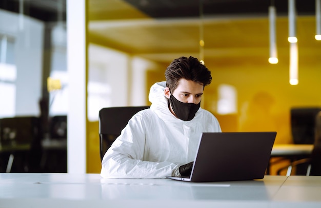 Человек в защитном костюме химзащиты и маске работает за компьютером в пустом офисе