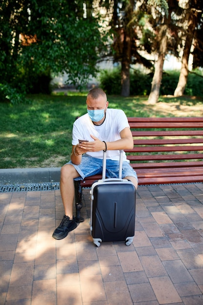 보호 마스크의 남자, 가방 및 휴대 전화, 코로나 바이러스 전염병 중 생활, 항공 여행 열기, 여행 개념과 함께 야외 공원에서 벤치에 앉아.