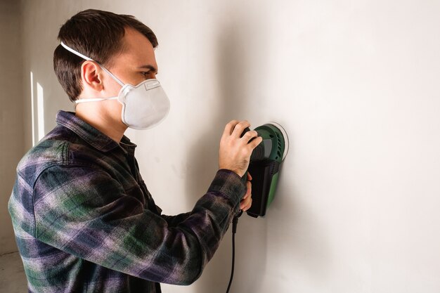 Uomo in maschera protettiva che lavora con levigatrice elettrica per levigare la superficie della parete in gesso, concetto di ristrutturazione della stanza