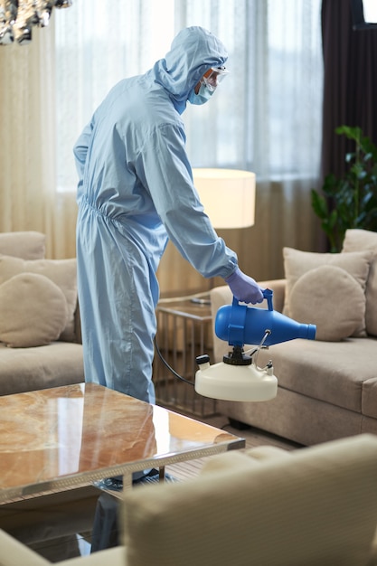 Man in protective overalls and a mask making sanitizing while standing in room. Coronavirus and quarantine concept