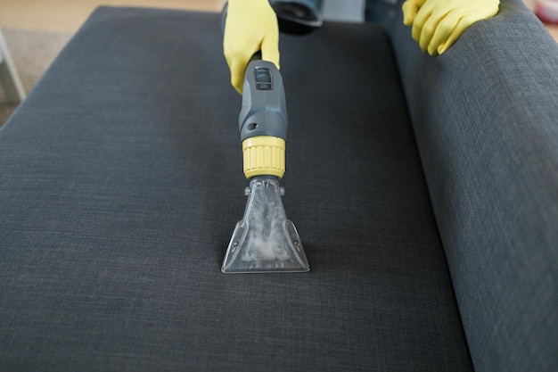 Человек в защитной резиновой перчатке чистит диван профессиональным методом извлечения с моющим пылесосом