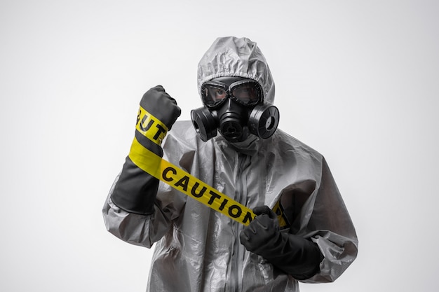 A man in a protective suit and gas mask holds a yellow tape "Caution" on  her hand on a white isolated background. Quarantine. Biological and chemical hazard.