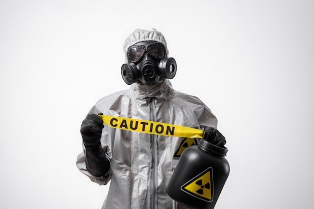 A man in a protective suit and gas mask holds a yellow tape "Caution" on a white isolated background. Quarantine. Biological and chemical hazard.