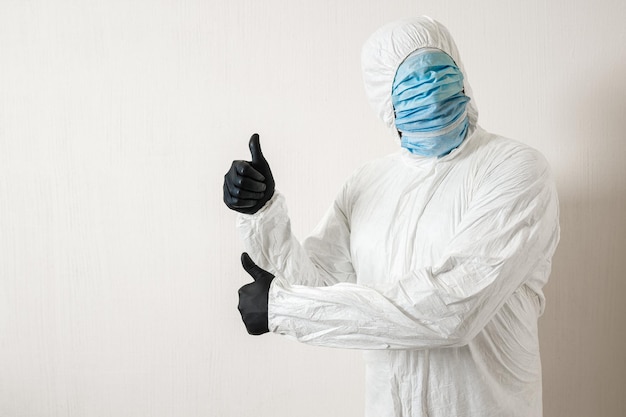A man in a protective suit hung with medical masks posing against a wall background showing various gestures with his fingers, the scientist shows a thumb up on both hands