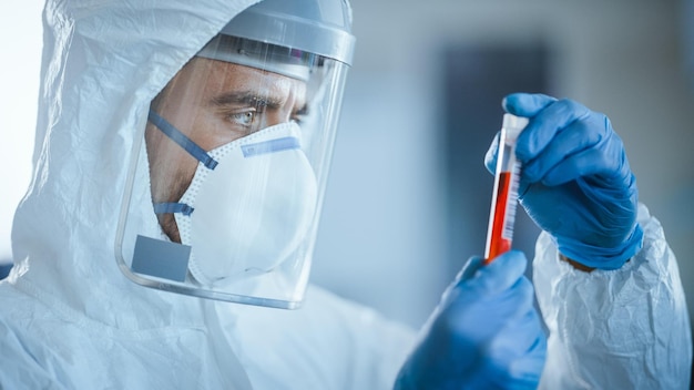 A man in a protective suit and mask holds a test tube of red liquid.