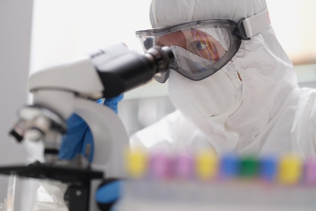 A man in a protective suit and mask looks through a microscope