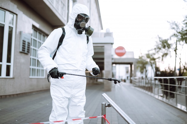 Man in protective suit and mask sprays disinfector onto the railing in the empty public place at dawn in the city of quarantine. Covid -19. Cleaning concept.