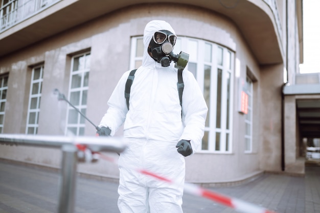 Man in protective suit and mask sprays disinfector onto the railing in the empty public place at dawn in the city of quarantine. Covid -19. Cleaning concept.