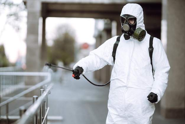 Человек в защитном костюме и маске распыляет дезинфектор на перила в пустом общественном месте на рассвете в городе Карантин. COVID-19. Концепция очистки.