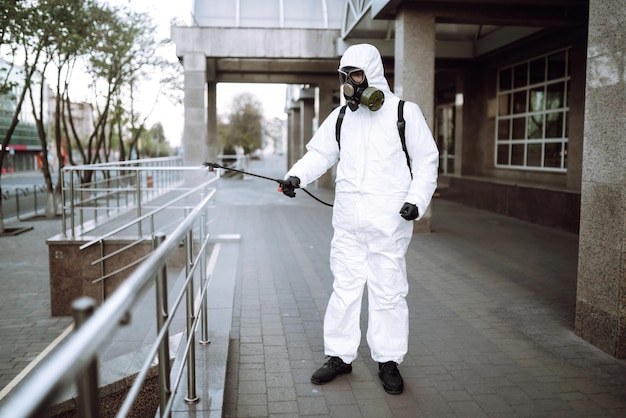 Man in protective suit and mask sprays disinfector onto the
railing in the public place covid 19