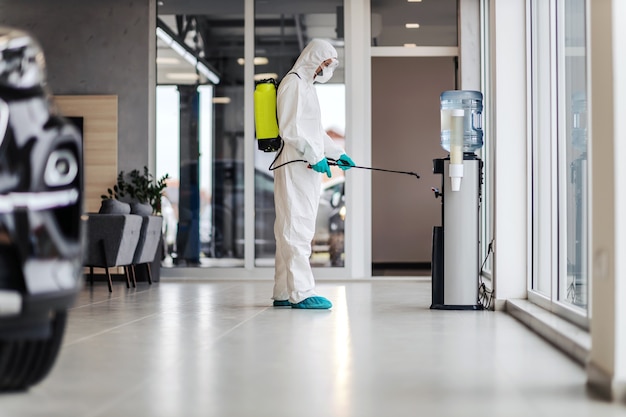 Foto uomo in uniforme protettiva con maschera e occhiali per disinfettare la macchina dell'acqua in un luogo moderno