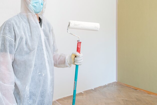 Man in protective work clothes painting the walls white colour with a paint roller.