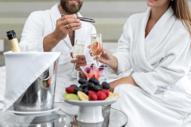 남자는 침대에 있는 호텔에서 그의 여자에게 스파클링 와인 한 잔에 딸기를 넣습니다