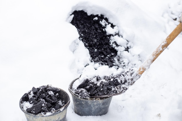 한 남자가 겨울 추운 눈 덮인 날에 삽으로 양동이에 석탄을 넣는다 눈 아래 석탄 더미 집 난방