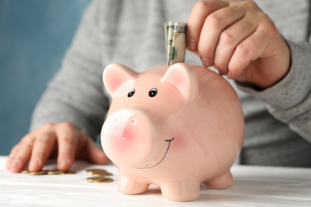 Man puts money in piggy bank on white wooden table, close up