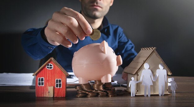 Man putting coin into piggy bank. Paper family and stack of coins on the table