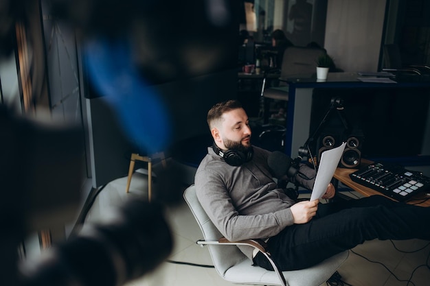 Man radio host or representer or journalist reads news from paper list in hand to studio microphone sitting at mixing sound panel or console at radio station toned