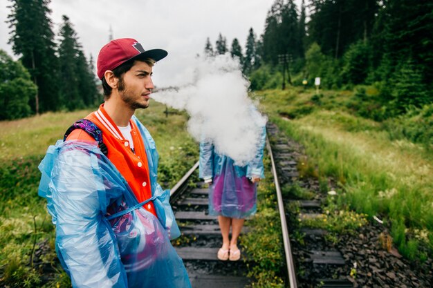 Man in raincoat blowing out clouds of smoke at railway