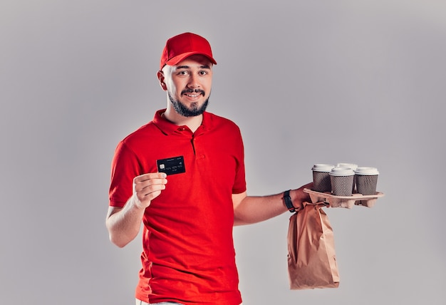 Man in red cap, t-shirt giving fast food order isolated on grey\
background. male courier holding credit card, paper packet with\
food, coffee. products delivery from shop or restaurant to\
home