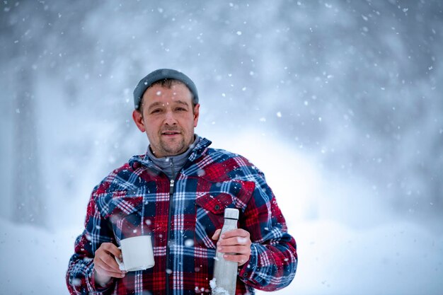 美しい雪の吹きだまりと森を背景に、降雪時に赤いチェックジャケットを着た男性がお茶を片手に持っています。