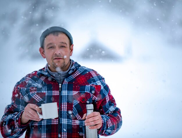 A man in a red check jacket is holding a cup of tea in his hands during a snowfall against the backdrop of beautiful snowdrifts and a forest