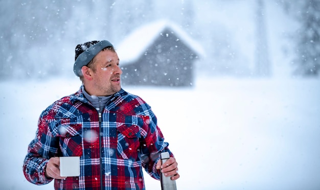 A man in a red check jacket is holding a cup of tea in his hands during a snowfall against the backdrop of beautiful snowdrifts and a forest