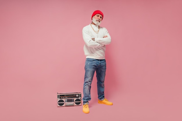 Man in red hat and jeans looking into camera on pink background Trendy guy with white beard in white sweatshirt posing