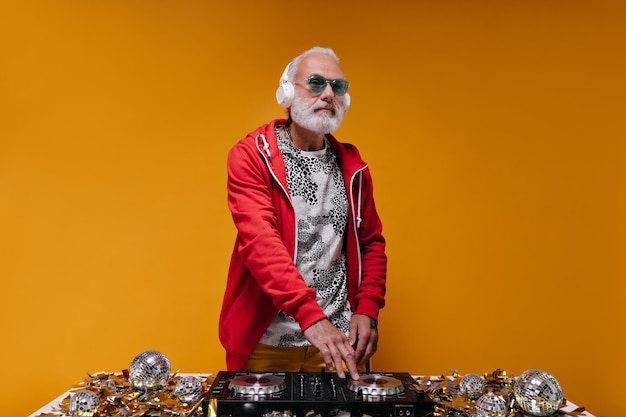 Man in red jacket and headphones makes music with Dj controller Adult guy with grey hair in stylish sweatshirt and blue sunglasses posing