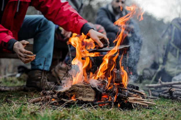 Man in a red jacket placing a cover on the black pan standing on the bonfire over the group of friends