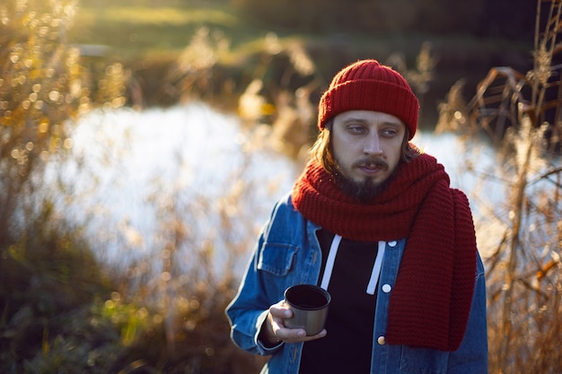 Man in a red knitted scarf and hat stands by lake in autumn and drinks tea mug thermos